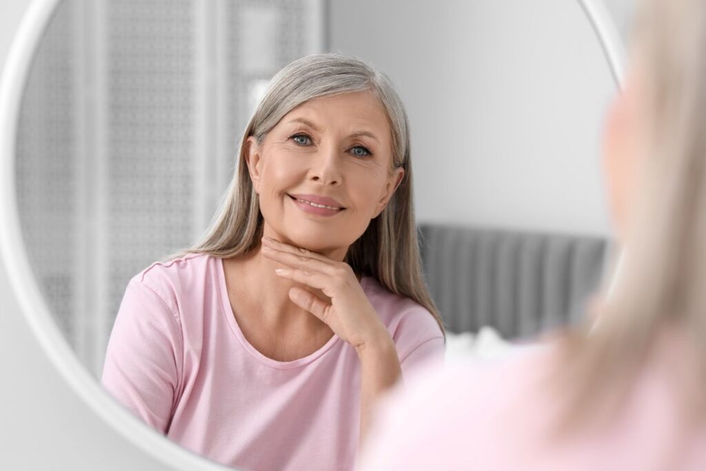 Ältere Frau mit grauen Haaren betrachtet ihr Gesicht im Spiegel – zufriedener Ausdruck dank natürlicher Anti-Aging-Routine
